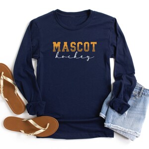May include: Navy blue long-sleeved shirt with the text "MASCOT hockey" in orange and white. The shirt is laid flat on a white surface with a pair of brown sandals and a pair of blue denim shorts.
