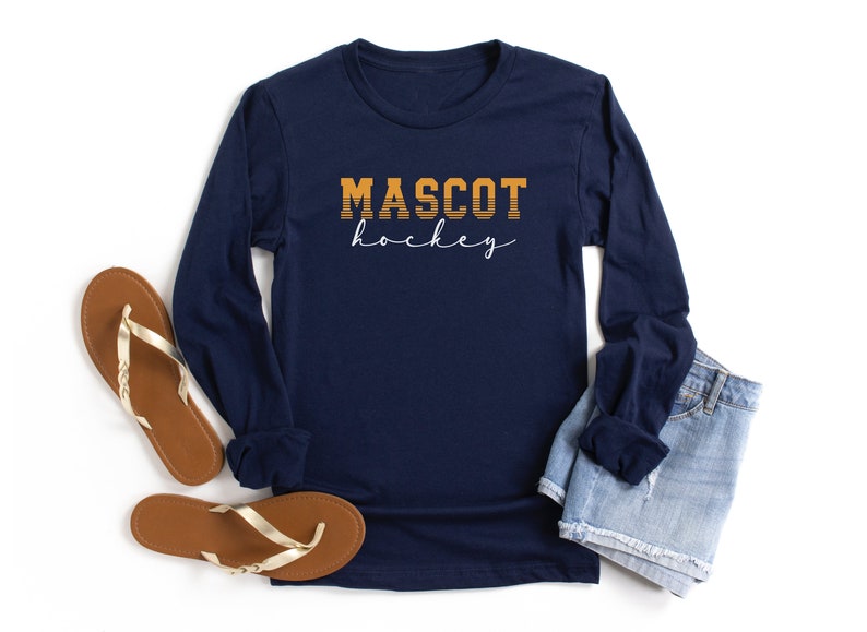 May include: Navy blue long-sleeved shirt with the text "MASCOT hockey" in orange and white. The shirt is laid flat on a white surface with a pair of brown sandals and a pair of blue denim shorts.