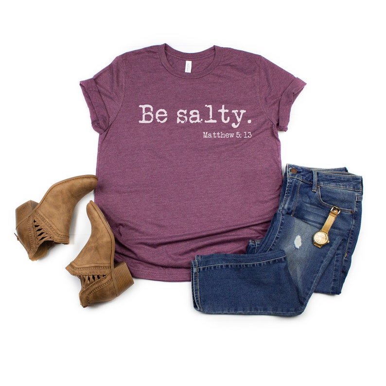 May include: A maroon t-shirt with the text "Be salty. Matthew 5:13" printed in white. The shirt is paired with blue jeans and brown ankle boots.