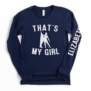 May include: A navy blue long-sleeved t-shirt with white text that reads "That's My Girl" and a silhouette of a woman doing a dance pose. The right sleeve has the name "Elizabeth" printed on it in white.