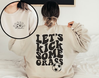 May include: A beige sweatshirt with the text "Let's Kick Some Grass" in black retro font on the back. A black and white soccer ball is below the text. The front of the sweatshirt has the text "Soccer Mom" in black with a small black and white soccer ball.