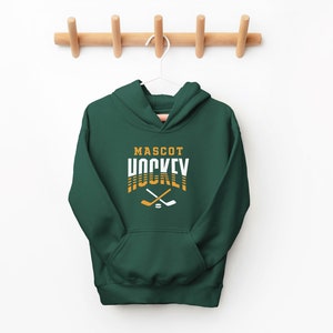 Puede incluir: Una sudadera con capucha verde oscuro con el texto "MASCOT HOCKEY" en letras blancas y doradas. Dos palos de hockey cruzados y un disco se encuentran debajo del texto.