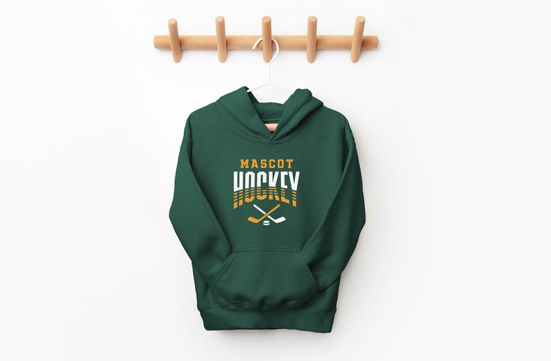 Puede incluir: Una sudadera con capucha verde oscuro con el texto "MASCOT HOCKEY" en letras blancas y doradas. Dos palos de hockey cruzados y un disco se encuentran debajo del texto.