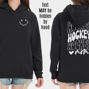 Peut inclure: Sweat-shirt &agrave; capuche noir avec un smiley blanc sur le devant et le texte "Hockey Mom" en police r&eacute;tro au dos.