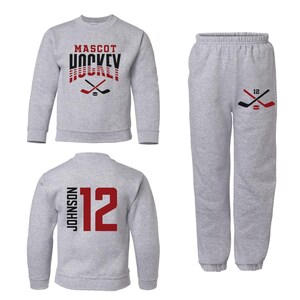 K&ouml;nnte beinhalten: Grauer Pullover mit dem Aufdruck "MASCOT HOCKEY" auf der Vorderseite und einer Grafik mit einem Hockeyschl&auml;ger. Eine passende graue Jogginghose mit einer Hockeyschl&auml;ger-Grafik und der Nummer "12" auf dem Bein. Ein zweiter grauer Pullover mit der Nummer "12" und dem Namen "JOHNSON" auf der R&uuml;ckseite.