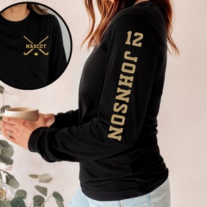 May include: Black long-sleeve shirt with a gold hockey stick graphic and the word "MASCOT" on the front. The shirt also has a gold number "12" and the name "JOHNSON" printed vertically on the left sleeve.