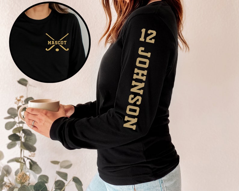 May include: Black long-sleeve shirt with a gold hockey stick graphic and the word "MASCOT" on the front. The shirt also has a gold number "12" and the name "JOHNSON" printed vertically on the left sleeve.