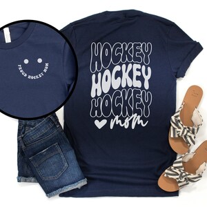 Peut inclure: T-shirt bleu marine avec un motif blanc qui dit "Hockey Hockey Hockey Mom" avec un c&oelig;ur. Le devant du t-shirt a un smiley blanc avec le texte "Proud Hockey Mom".