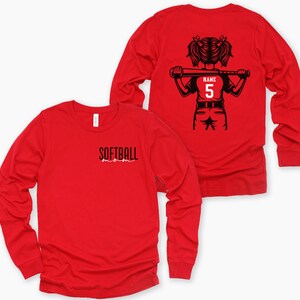 Puede incluir: Camiseta de softbol roja de manga larga. La parte delantera de la camiseta tiene la palabra "SOFTBALL" impresa en negro. La parte trasera de la camiseta presenta un gr&aacute;fico negro de una ni&ntilde;a sosteniendo un bate, con el n&uacute;mero "5" en la camiseta. La palabra "NAME" est&aacute; encima del n&uacute;mero.