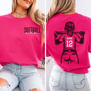 May include: Hot pink sweatshirt with the text "SOFTBALL mom" on the front. The back features a black graphic of a softball player holding a bat, with space for a name and the number 12. The sweatshirt is paired with light blue jeans.