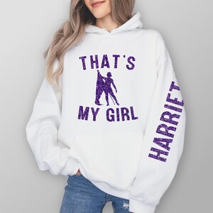 May include: White hoodie with purple glitter text that says "That's My Girl" and "Harriett" on the sleeve.