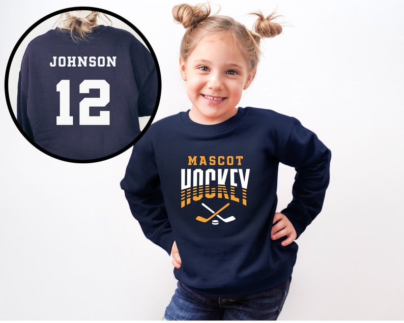 Puede incluir: Sudadera azul marino con el texto "MASCOT HOCKEY" en naranja y blanco con palos de hockey cruzados y un disco. La parte posterior de la sudadera tiene el texto "JOHNSON 12" en blanco.
