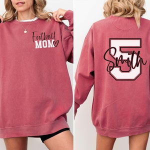 K&ouml;nnte beinhalten: Ein maroonfarbener Sweatshirt mit dem Text "Football Mom" und einem Herz auf der Vorderseite und einer wei&szlig;en Nummer 5 mit dem Text "Smith" auf der R&uuml;ckseite.