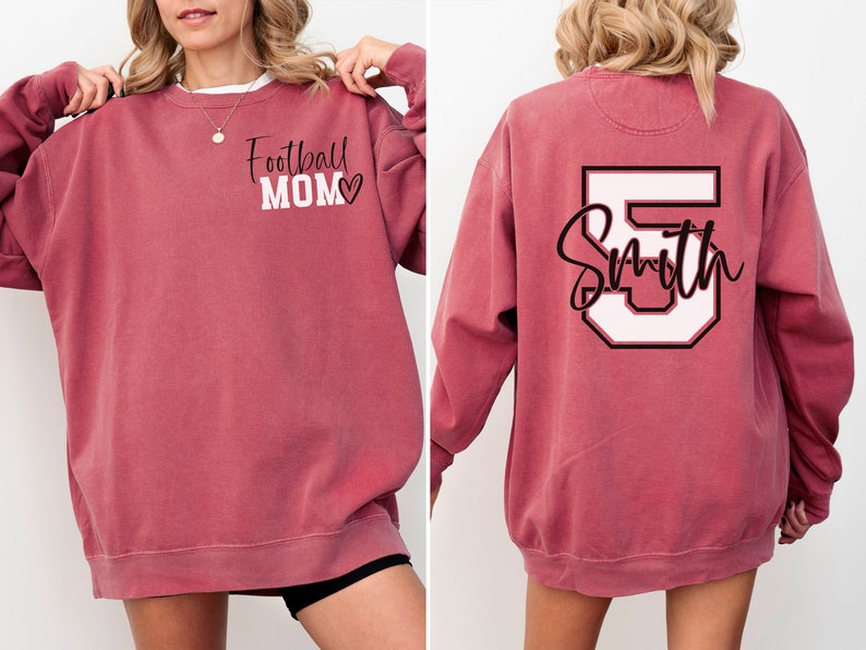 K&ouml;nnte beinhalten: Ein maroonfarbener Sweatshirt mit dem Text "Football Mom" und einem Herz auf der Vorderseite und einer wei&szlig;en Nummer 5 mit dem Text "Smith" auf der R&uuml;ckseite.