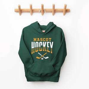 K&ouml;nnte beinhalten: Ein dunkelgr&uuml;ner Kapuzenpullover mit dem Text "MASCOT HOCKEY" in wei&szlig;er und goldener Schrift. Der Text ist in einer Retro-Schriftart gehalten. Zwei gekreuzte Hockeyschl&auml;ger und ein Puck befinden sich unter dem Text.