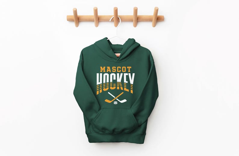 K&ouml;nnte beinhalten: Ein dunkelgr&uuml;ner Kapuzenpullover mit dem Text "MASCOT HOCKEY" in wei&szlig;er und goldener Schrift. Der Text ist in einer Retro-Schriftart gehalten. Zwei gekreuzte Hockeyschl&auml;ger und ein Puck befinden sich unter dem Text.