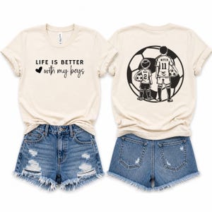 May include: A cream-colored t-shirt featuring the black text 'LIFE IS BETTER with my boys' and a heart icon on the front. The back displays a large black graphic of a soccer ball with two figures in soccer uniforms labeled 'MAX 2' and 'MITCH 11'.