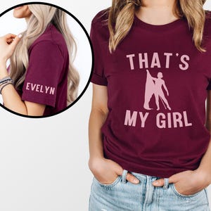 May include: Burgundy t-shirt with the text "THAT'S MY GIRL" in pink lettering and a silhouette of a figure holding a baton. The sleeve has the name "EVELYN" in pink lettering.