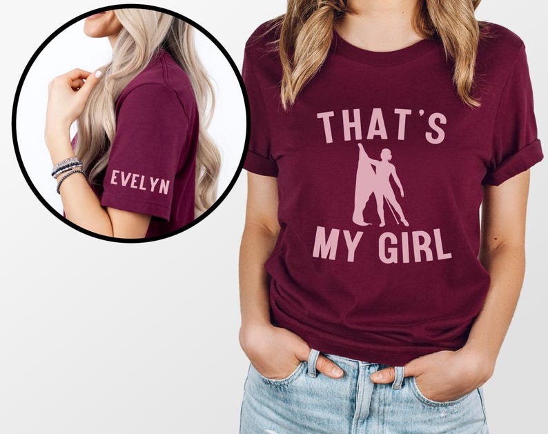 May include: Burgundy t-shirt with the text "THAT'S MY GIRL" in pink lettering and a silhouette of a figure holding a baton. The sleeve has the name "EVELYN" in pink lettering.