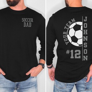 May include: Black long-sleeve shirt with a soccer ball graphic and the text "Your Team #12 JOHNSON" on the back. The front of the shirt says "Soccer Dad".