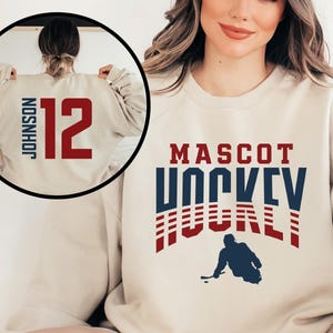 May include: A cream-colored sweatshirt with a blue and red graphic that says "MASCOT HOCKEY" with a hockey player silhouette. The back of the sweatshirt has a red number "12" and the name "JOHNSON" in black.