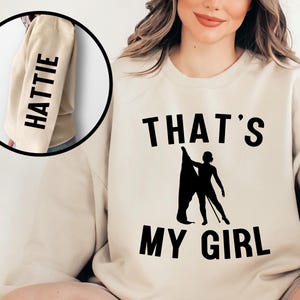 May include: A beige sweatshirt with the text "THAT'S MY GIRL" and a silhouette of a person holding a flag. The left sleeve has the name "HATTIE" printed vertically.