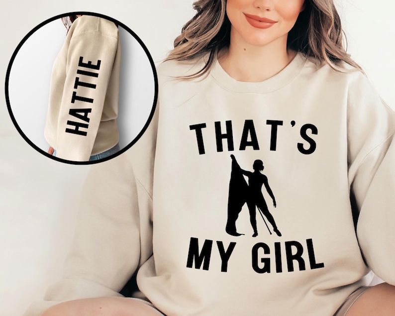 May include: A beige sweatshirt with the text "THAT'S MY GIRL" and a silhouette of a person holding a flag. The left sleeve has the name "HATTIE" printed vertically.