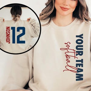 May include: A beige sweatshirt with the text "YOUR TEAM softball" in blue and red on the right side. The back of the sweatshirt has the number "12" in blue with the name "JOHNSON" in blue above it.