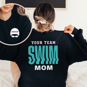 Peut inclure: Sweat-shirt noir avec le texte "Your Team Swim Mom" en lettres turquoise et blanches. Une petite image d'un bonnet de bain avec le texte "Johnson" est sur le devant du sweat-shirt.