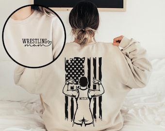 Puede incluir: Una sudadera beige con el texto "Wrestling Mom" y un corazón en la parte delantera. La parte posterior de la sudadera presenta una ilustración en blanco y negro de un luchador frente a una bandera estadounidense con el nombre "Mitch" impreso debajo.
