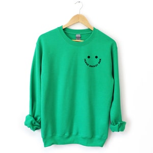 Peut inclure: Sweat-shirt vert avec un smiley noir et le texte "Proud Hockey Mom" sur le devant.