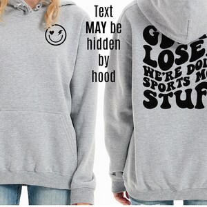 K&ouml;nnte beinhalten: Grauer Kapuzenpullover mit einem schwarzen Smiley-Gesicht mit einem Herz und einem Blitz auf der Vorderseite und dem Text "GIVEN LOSER WE'RE DOING SPORTS MOM STUFF" auf der R&uuml;ckseite.