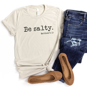 May include: A beige t-shirt with the text "Be salty. Matthew 5:13" printed on the front. The shirt is paired with blue jeans and brown sandals.