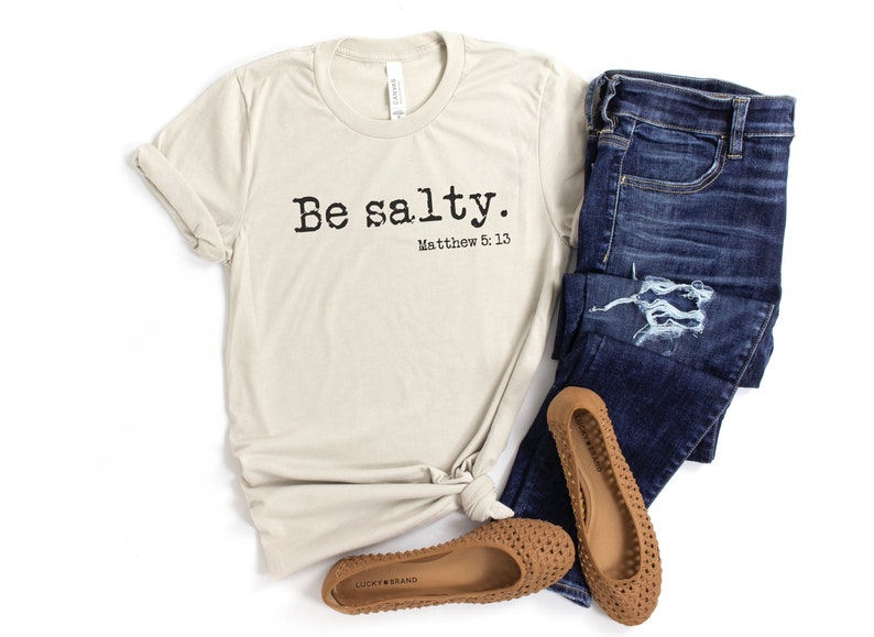 May include: A beige t-shirt with the text "Be salty. Matthew 5:13" printed on the front. The shirt is paired with blue jeans and brown sandals.