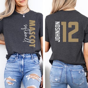 May include: A dark gray t-shirt with a gold and white design. The front of the shirt says "MASCOT softball" and the back of the shirt says "JOHNSON 12".