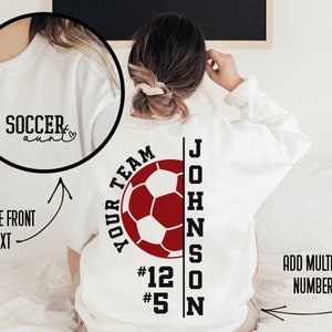 May include: White sweatshirt with a red soccer ball graphic and the text "YOUR TEAM" above the ball. The text "JOHNSON" is vertically aligned to the right of the ball with the numbers "#12" and "#5" below the text. The text "SOCCER aunt" is on the front of the sweatshirt.