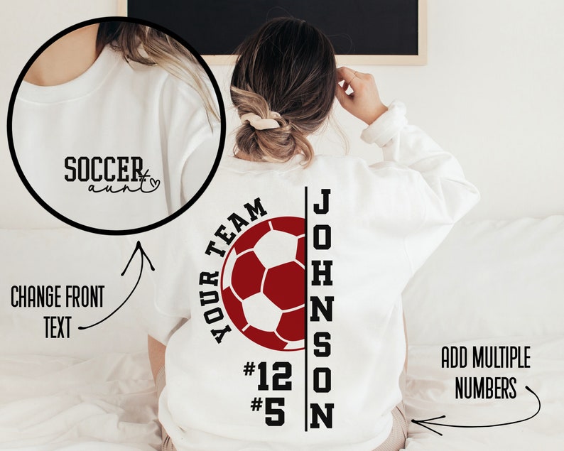 May include: White sweatshirt with a red soccer ball graphic and the text "YOUR TEAM" above the ball. The text "JOHNSON" is vertically aligned to the right of the ball with the numbers "#12" and "#5" below the text. The text "SOCCER aunt" is on the front of the sweatshirt.