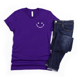 Peut inclure: T-shirt &agrave; manches courtes de couleur violette avec un smiley blanc et le texte "Proud Hockey Mom" autour.