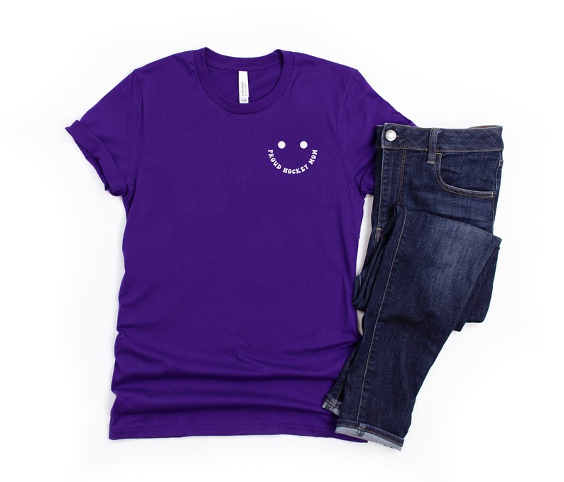 Peut inclure: T-shirt &agrave; manches courtes de couleur violette avec un smiley blanc et le texte "Proud Hockey Mom" autour.