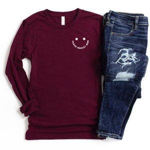Peut inclure: Un t-shirt &agrave; manches longues bordeaux avec un smiley blanc qui dit "Good Hockey Mom". Le t-shirt est associ&eacute; &agrave; un jean bleu.