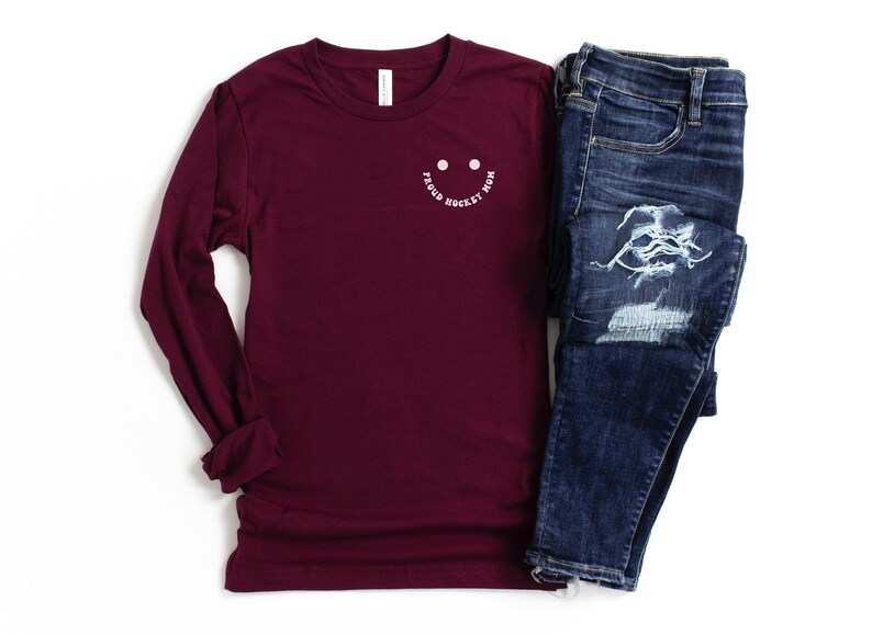 Peut inclure: Un t-shirt &agrave; manches longues bordeaux avec un smiley blanc qui dit "Good Hockey Mom". Le t-shirt est associ&eacute; &agrave; un jean bleu.