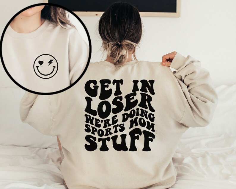 K&ouml;nnte beinhalten: Ein beiger Pullover mit einem schwarzen Aufdruck auf der R&uuml;ckseite, der in einer Retro-Schrift "Get in Loser We're Doing Sports Mom Stuff" lautet. Die Vorderseite des Pullovers hat ein kleines schwarzes Smiley mit einem Herz und einem Blitz.