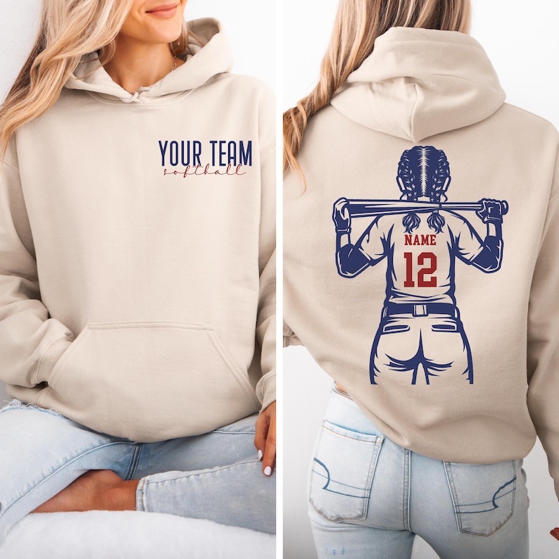 Puede incluir: Sudadera con capucha beige con bolsillo delantero y capucha con cord&oacute;n. La parte delantera presenta el texto "YOUR TEAM softball" en azul marino y rojo. La parte trasera muestra un gr&aacute;fico de jugadora de softbol con un bate, el nombre y el n&uacute;mero 12 en rojo.