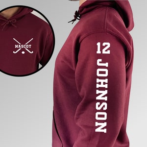 May include: Burgundy hooded sweatshirt with white text on the sleeve that reads "12 JOHNSON". The front of the sweatshirt has a white graphic of two crossed hockey sticks with a puck in the middle and the word "MASCOT" underneath.