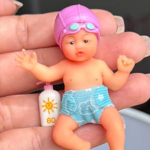 May include: A miniature baby doll wearing a pink swim cap with light blue goggles and a light blue patterned diaper. The doll is accompanied by a small white bottle with a pump dispenser, featuring a yellow sun graphic and the number 60 printed on it.