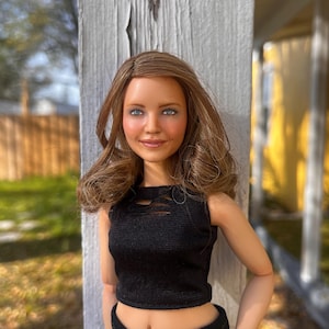 May include: A doll with shoulder-length brown hair and blue eyes, wearing a black sleeveless crop top with a distressed neckline and matching black pants. The doll has a fair complexion, a soft smile, and is positioned against a vertical wooden post.