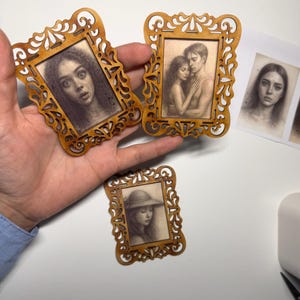 May include: Miniature wooden picture frames with intricate cut-out designs. Each frame holds a pencil sketch portrait. The frames are a warm brown color, and the portraits depict people. The frames are held in a hand.
