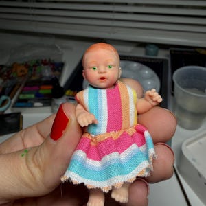 May include: A miniature doll with a fair complexion, red hair, and green eyes. The doll wears a colorful striped dress with pink, blue, and orange hues. The doll is approximately 3 inches tall, and is a collectible item.