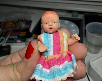 May include: A miniature doll with a fair complexion, red hair, and green eyes. The doll wears a colorful striped dress with pink, blue, and orange hues. The doll is approximately 3 inches tall, and is a collectible item.