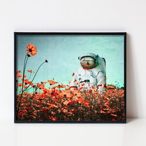K&ouml;nnte beinhalten: Ein Astronaut in einem wei&szlig;en Raumanzug steht in einem Feld mit orangefarbenen Kosmosblumen vor einem blauen und gr&uuml;nen Himmel.